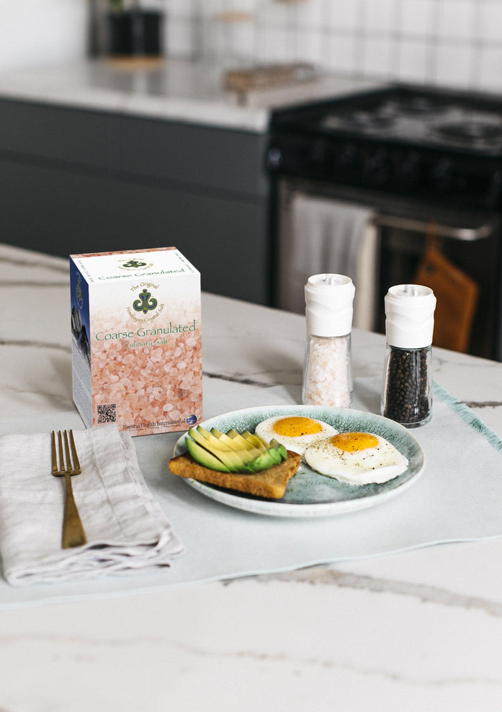 Coarse Granulated Culinary Salt right-facing front of Coarse Granulated Culinary Salt product box showing salt granules with salt and spoon, on white and black marble kitchen counter with grinder filled with black pepper and grinder filled with salt, gold fork on white cloth napkin, and plate of avocado toast and sunny-side-up eggs