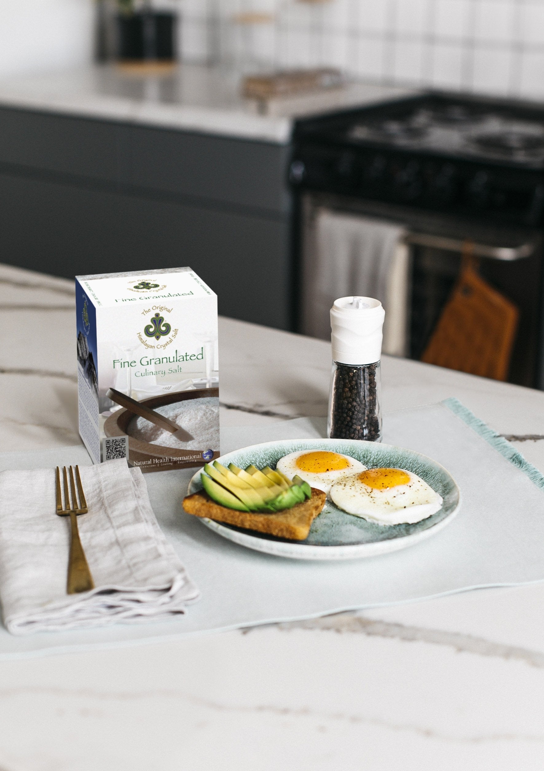 Fine Granulated Culinary Salt right-facing front of Finely Granulated Culinary Salt product box showing wooden bowl with salt and spoon, on white and black marble kitchen counter with grinder filled with black pepper, gold fork on white cloth napkin, and plate of avocado toast and sunny-side-up eggs