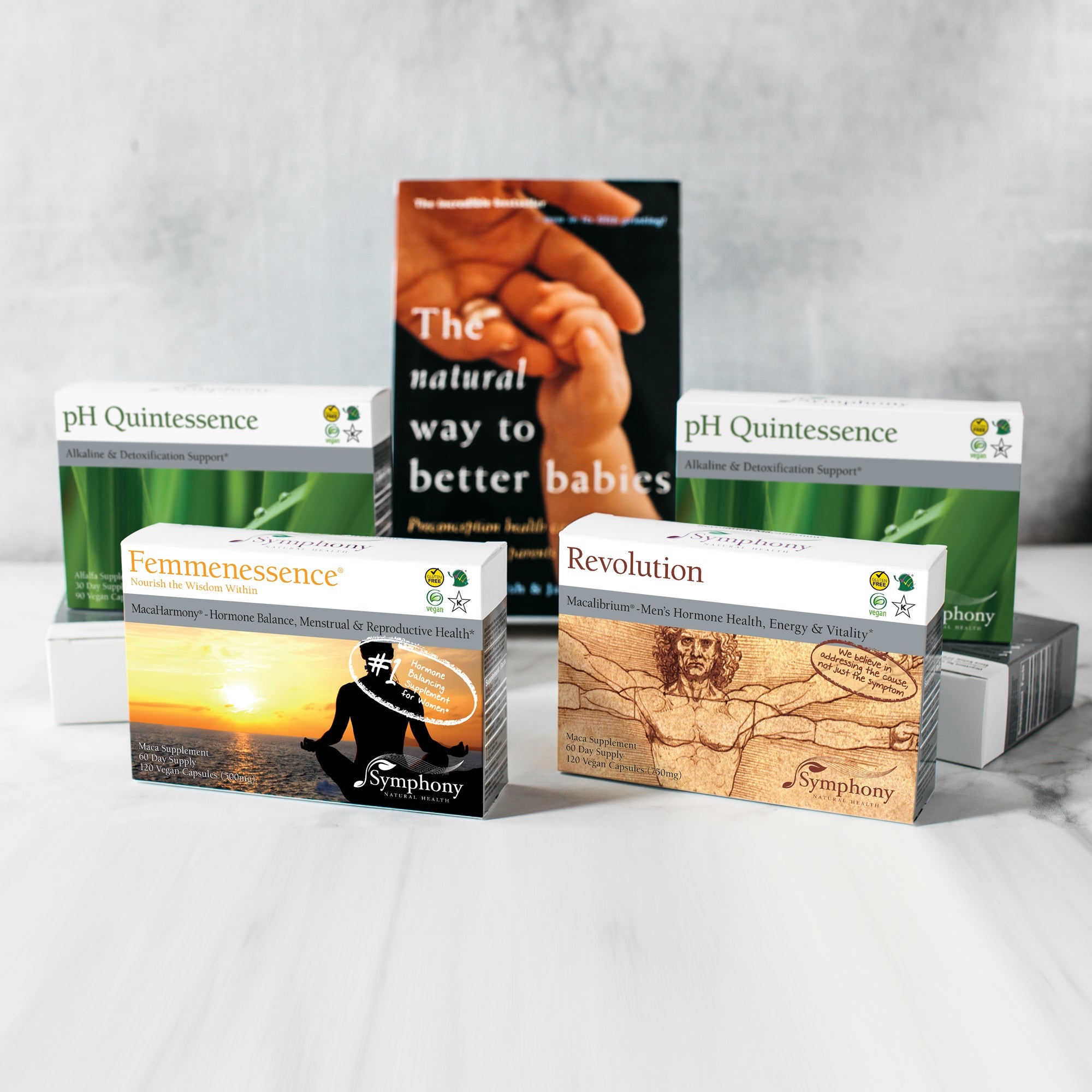 Fertility Preconception Pack one MacaHarmony boxes showing silhouette of woman facing horizon at sunset, one Revolution Macalibrium with Vitruvian Man illustration, we believe in addressing the cause not just the symptom, two ph Quintessence with green leaves with water drops, with leaning book The Natural Way to Better Babies, vegan, gluten free, Kosher, on gray background