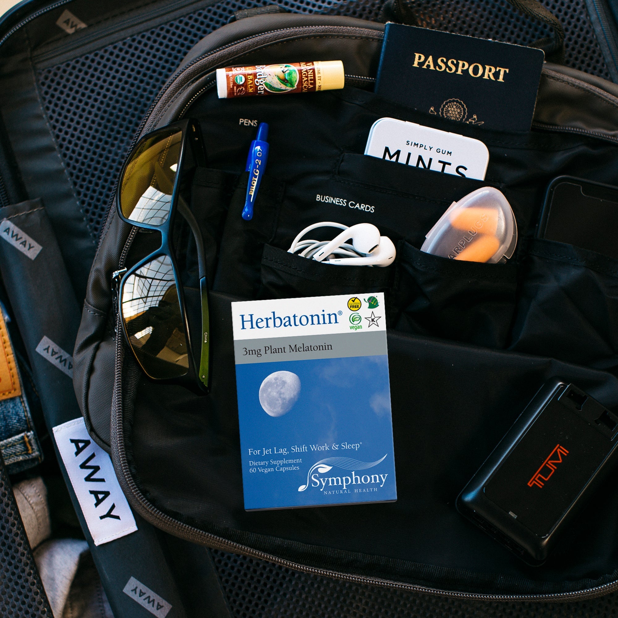 Away-brand black suitcase opened with contents of sunglasses, mints tin, passport book, lip balm, ear pods, orange ear plugs, spa wipes, Femmenessence MacaHarmony box, and Herbatonin Travel Pack box in woman's hand