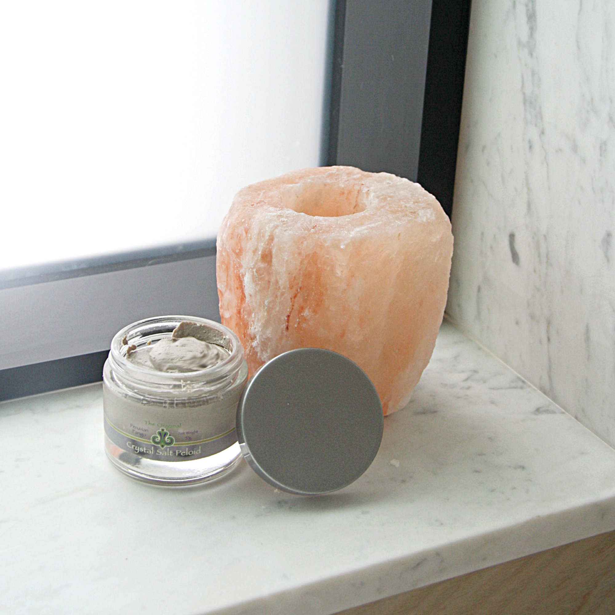 Sole Peloid Mud Mask glass jar with leaning silver lid and Himalayan Crystal Salt logo on window sill with salt votive