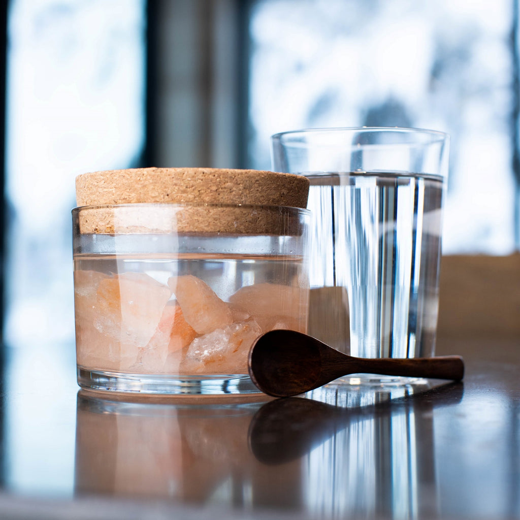 Sole Jar Plus Stones product glass jar filled with Himalayan Crystal Salt stones filled and water, with cork cover,  glass of water and woodens spoon resting on table with snowy window in background