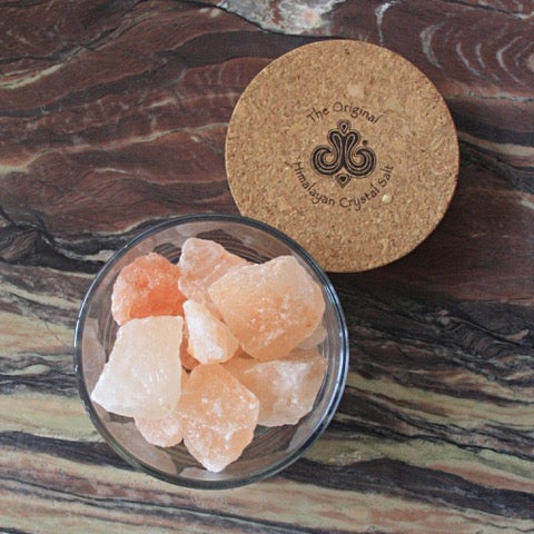 Sole Jar Plus Stones product glass jar filled with Himalayan Crystal Salt stones with cork cover featuring Himalayan Crystal Salt logo both resting on dark wood surface