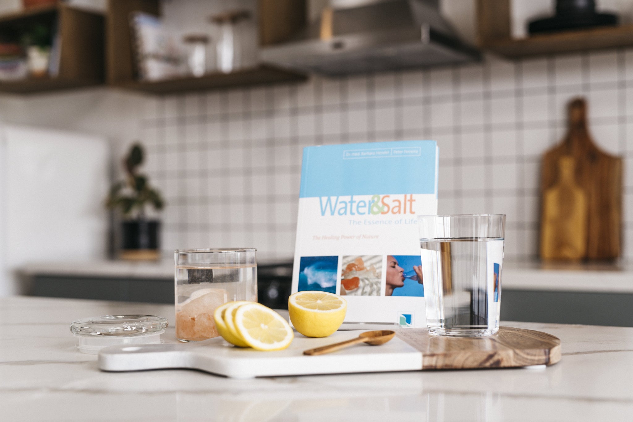 Turquoise and white Water & Salt The Essence of Life Book showing blue ocean, salt stones and woman drinking water from clear bottle, leaning on kitchen counter with glass jar filled with Himalayan Crystal Salt Stones and water, sliced lemons, wooden spoon, glass of water and cutting board