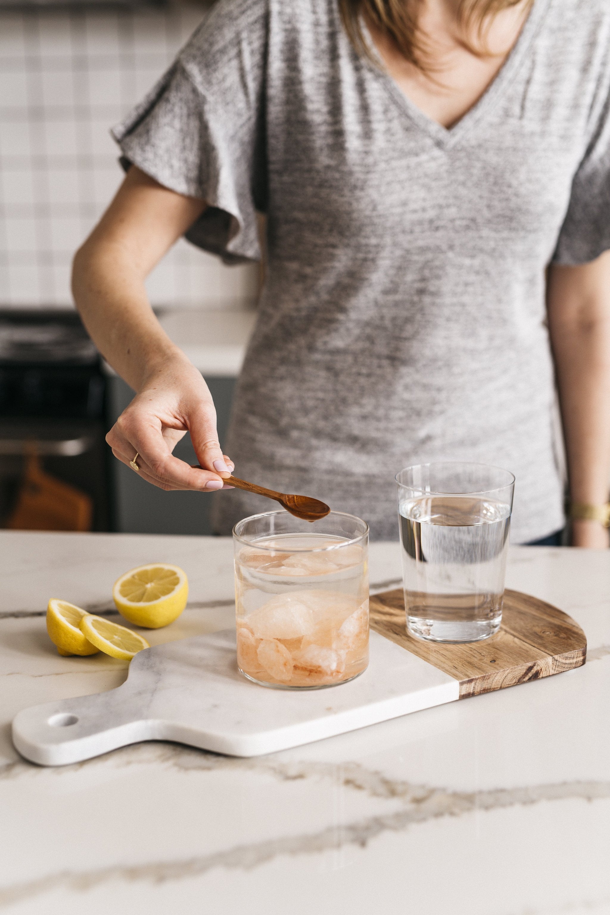 Woman in gray t-shirt holding wooden spoon over glass jar filled with Himalayan Crystal Salt Stones and water, with sliced lemons, glass of water and cutting board on light gray and white marble kitchen counter 